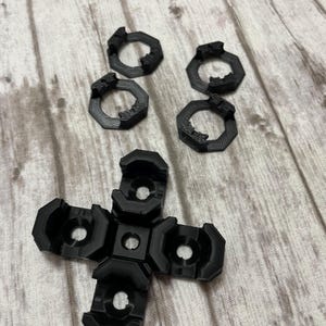 May include: Black 3D-printed replacement parts for a gaming controller. The image shows a cross-shaped component and four circular components. The parts are arranged on a light-colored, wood-grain surface.