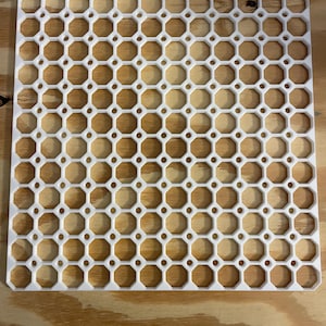 Może przedstawiać: Kwadratowa, biała plastikowa kratka z powtarzającym się wzorem ośmiokątnych otworów. Każdy otwór zawiera drewnianą, ośmiokątną wkładkę. Drewniane wkładki różnią się kolorem od jasnobrązowego do ciemnobrązowego.