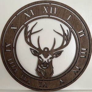 May include: A round, wooden wall clock with a deer head silhouette at the center. The clock face features Roman numerals and a dark brown finish. The deer head is intricately cut out, adding depth to the design.