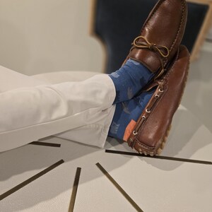 May include: Brown leather driving shoes with a decorative bow detail. The shoes are worn with white trousers and colourful socks.