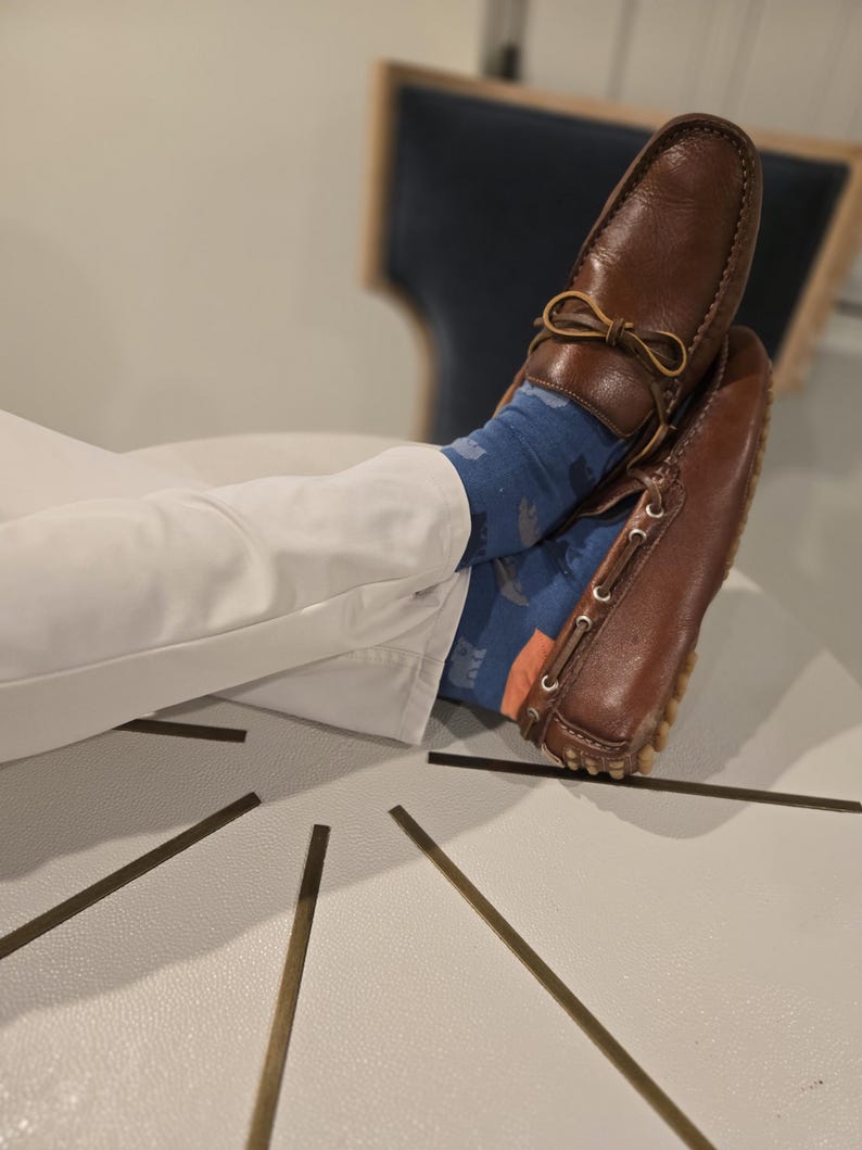 May include: Brown leather driving shoes with a decorative bow detail. The shoes are worn with white trousers and colourful socks.