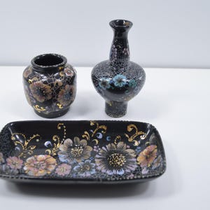 May include: A set of three black ceramic decorative items: a small vase, a taller vase, and a rectangular dish. Each piece is adorned with floral patterns in gold, pink, and blue hues. The items are arranged on a white surface.