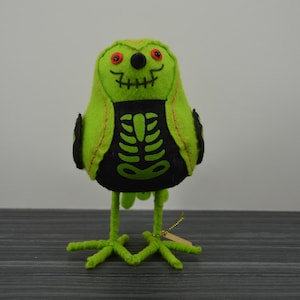 May include: A lime green felt bird decoration with a black skeleton design on its chest. The bird has red eyes, a black beak, and a stitched mouth. It stands on green wire legs and feet, perfect for Halloween or spooky decor.