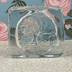 May include: A clear, square glass paperweight with a detailed embossed image of a cat. The cat is curled up, with its head resting on its paws. The paperweight is sitting on a speckled surface.
