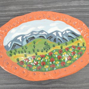 May include: Oval ceramic plate with a hand-painted mountain landscape. The plate features a vibrant orange border with a textured design. The central image depicts snow-capped mountains, a green hillside, and colorful flowers.