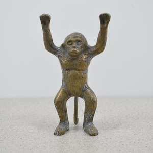 May include: A bronze-colored monkey figurine with its arms raised. The monkey is standing upright with its legs slightly apart and a long tail. The figurine has detailed facial features and a textured surface, giving it a realistic appearance.