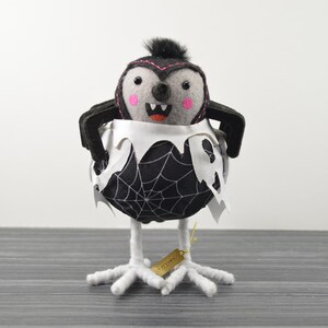May include: A Halloween-themed felt bird with a spider web design. The bird has a gray face with pink cheeks, black wings, and a black mohawk. It is perched on white, claw-like feet. A small tag hangs from its feet.