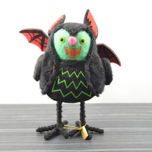 May include: A whimsical Halloween decoration featuring a black bat-like creature with a green face, red wings, and a zig-zag pattern on its chest. The bat has a pink nose and is perched on spindly legs.