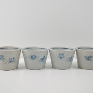 May include: Four small, off-white ceramic cups with a blue floral design. Each cup has a slightly irregular shape and a thin blue line around the rim. The cups are arranged in a row against a white background.