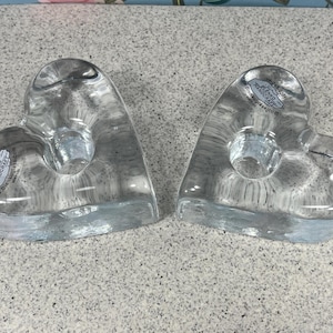 May include: Two clear glass heart-shaped candle holders. Each has a circular indentation for a candle and a sticker with text. The candle holders are made of transparent glass and are displayed on a speckled surface.