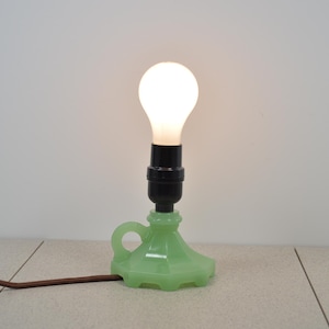 May include: A small, green glass lamp with a black socket and a lit, round light bulb. The lamp has a handle and a decorative base. A brown electrical cord extends from the lamp.