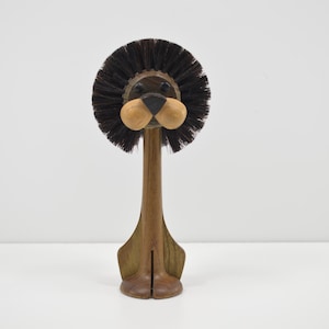May include: A wooden lion-shaped brush with a dark brown bristle mane and a light brown nose. The brush has a long, slender handle and a wide base. The lion's face is detailed with black eyes and a black nose.