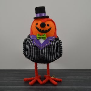 May include: A Halloween-themed decorative figure of a bird with an orange head and a carved jack-o'-lantern face. It wears a black and white pinstriped suit with a purple lapel and a green bow tie. The bird has red feet and a black top hat.