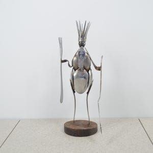 May include: A whimsical metal sculpture of a figure made from spoons, forks, and other utensils. The sculpture stands on a round wooden base and holds two forks. The figure has a crown-like structure on its head.