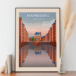 Op de afbeelding: Een vintage reisposter met een kleurrijke illustratie van de Speicherstadt-wijk in Hamburg, Duitsland. De poster toont een kanaal met rode bakstenen gebouwen aan weerszijden en een groot gebouw in het midden. De tekst "HAMBURG GERMANY" staat bovenaan de poster.