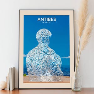 Peut inclure: Une affiche avec un fond bleu et une sculpture blanche d'une personne faite de lettres. L'affiche indique "ANTIBES FRANCE" en haut.