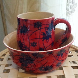 May include: A red ceramic mug with a handle and a matching bowl. Both items feature a blue floral design. The mug sits inside the bowl. The interior of both pieces is cream-coloured.