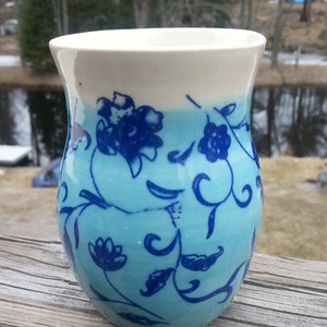 May include: A white ceramic mug with a blue and white floral design. The mug has a light blue glaze with a white rim. The floral design is a repeating pattern of blue flowers and leaves.