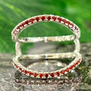 May include: A silver ring with two bands, each lined with small, red gemstones. The ring is set against a blurred background of green leaves and a reflective, gray surface. The ring's design is modern and elegant.