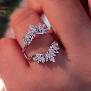 May include: A silver ring with a crown-like design, adorned with multiple clear, sparkling gemstones. The ring is held up by a hand, showcasing the intricate details of the jewelry. The gemstones are arranged in a leaf-like pattern.