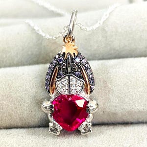 May include: A silver necklace with a pendant in the shape of a cartoon character. The pendant has a red heart-shaped gem, a silver frame, and purple and white detailing. The necklace is presented on a light grey surface.