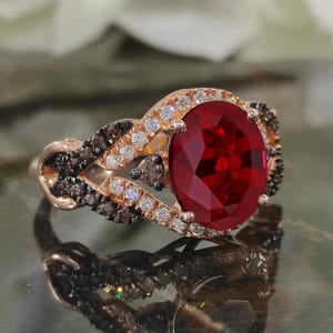 May include: Rose gold ring featuring a large, oval-cut, deep red gemstone as the centerpiece. The ring is accented with smaller white and dark brown stones, set in a braided design around the main stone. The ring's band has a twisted, intricate pattern.