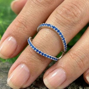 Peut inclure: Une bague en argent avec un design à double anneau, ornée de petites pierres précieuses bleues vives. La bague est présentée sur un doigt, mettant en valeur ses détails complexes et son aspect scintillant. Les pierres sont espacées uniformément.