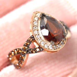 Può includere: Anello d'oro con una gemma rosso scuro a forma di pera. La gemma è circondata da piccole pietre chiare. La fascia presenta un design a infinito con pietre marroni e nere, su sfondo rosa.