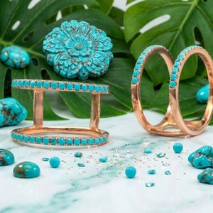May include: Rose gold rings and a double-band ring are adorned with turquoise stones. A carved turquoise flower and loose turquoise stones are scattered on a marble surface, with a backdrop of green leaves.
