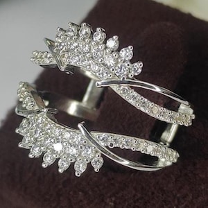 May include: A silver ring with a double band design. The ring features two curved bands, each adorned with small, sparkling clear stones. The top of the ring has a decorative, leaf-like design, also set with clear stones. The ring is displayed on a brown surface.