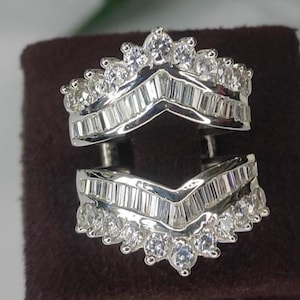 May include: Two-piece silver ring guard with a V-shaped design. The rings are encrusted with multiple small, clear, round and baguette-cut stones. The rings are displayed on a dark brown surface.