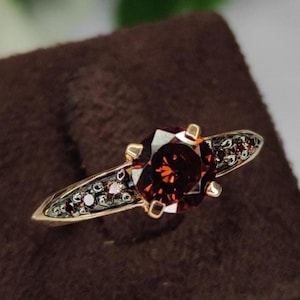 Può includere: Un anello d'oro con una grande pietra preziosa rosso scuro sfaccettata al centro. La fascia è adornata con pietre scure più piccole. L'anello è esposto su una superficie marrone vellutata, con uno sfondo sfocato bianco e verde.