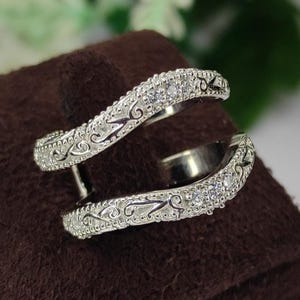 Puede incluir: Un par de realzadores de anillos plateados, curvos, con un diseño decorativo y pequeñas piedras preciosas transparentes. Los realzadores se exhiben sobre una superficie de terciopelo marrón, con un fondo borroso de follaje blanco y verde.