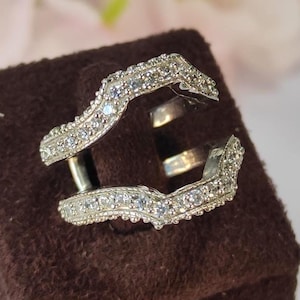 May include: A silver ring guard with a curved design, encrusted with small, sparkling stones. The ring guard is displayed on a brown velvet stand, with a blurred floral background.