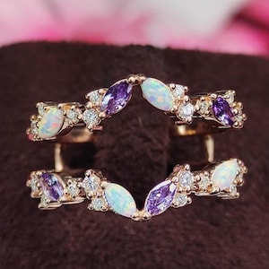 Op de afbeelding: Een goudkleurige ringversterker met een gebogen ontwerp. Het heeft afwisselende marquise-geslepen paarse en witte edelstenen, samen met kleine heldere accentstenen. De ring wordt weergegeven op een bruin oppervlak met een vage roze bloemenachtergrond.