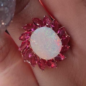 May include: A gold ring featuring a large, oval, white opal gemstone surrounded by smaller, pink gemstones. The ring is set against a blurred background of green and silver holiday decorations.
