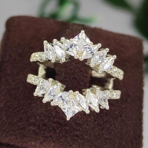 Puede incluir: Un anillo plateado con un diseño de doble banda. El anillo presenta una serie de piedras claras, de corte cuadrado y marquesa, engastadas en un patrón de halo. El anillo se exhibe sobre una superficie de terciopelo marrón, con follaje verde en el fondo.