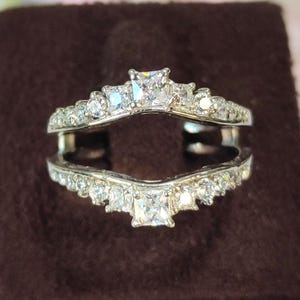 May include: A silver ring guard with a double band design. The ring features multiple small, clear gemstones and a larger square gemstone at the center of each band. The ring is displayed on a brown surface.