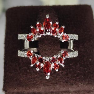 Pode incluir: Uma proteção de anel de prata com um halo de pedras preciosas vermelhas. A proteção do anel apresenta um design de banda dupla com uma abertura central. As pedras preciosas têm formato de marquesa e são dispostas em um padrão floral.