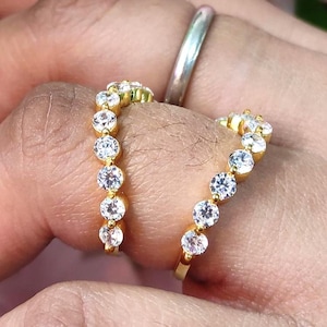 May include: Two gold-colored rings with clear, round gemstones. One ring is on a finger, and the other is held in a hand. A silver ring is also visible on a finger. The background features pink and green floral elements.