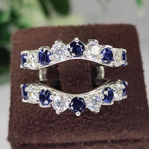 May include: Two silver ring enhancers, each featuring a curved design. The rings are adorned with alternating round, dark blue and clear gemstones. The enhancers are displayed on a brown stand, with a blurred background of green foliage.