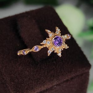 Puede incluir: Un anillo dorado con un diseño de sol, una piedra preciosa púrpura central y piedras blancas. El anillo tiene una banda delicada con piedras púrpuras. El anillo se muestra sobre una superficie marrón.