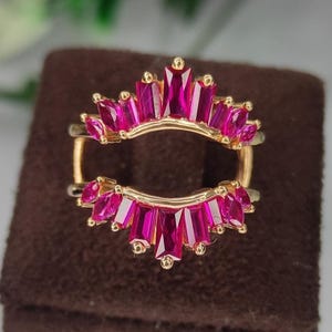 Może przedstawiać: Złoty pierścionek o podwójnym wzorze, ozdobiony żywymi różowymi kamieniami szlachetnymi. Kamienie szlachetne są cięte w bagietki i ułożone w wzór promieni słonecznych, tworząc olśniewający pokaz koloru i światła. Pierścionek jest umieszczony na brązowym tle.