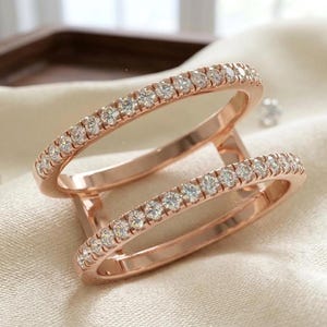 Peut inclure: Une bague double en or rose composée de deux anneaux parallèles sertis de petites pierres rondes transparentes sur le dessus. Les deux anneaux sont reliés par un pont métallique vertical, formant un bijou unique et cohérent.