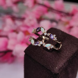 Op de afbeelding: Een goudkleurige ring met twee banden, versierd met ovale opalen, paarse edelstenen en kleine, heldere kristallen. De ring wordt gepresenteerd op een donkerbruin oppervlak, met een vage roze bloemenachtergrond.