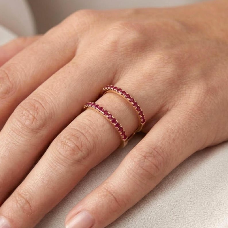 Pu&ograve; includere: Un anello a doppia fascia con una fascia dorata e una fila di piccole pietre preziose rosse su ogni fascia. L'anello &egrave; indossato su un dito, mettendo in mostra il design e il colore del gioiello.