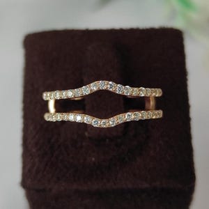 Op de afbeelding: Roségouden ringbeschermer met een dubbel bandontwerp. Elke band is ingelegd met kleine, sprankelende diamanten. De ring staat tegen een donkerbruine achtergrond, wat de details en schittering benadrukt.