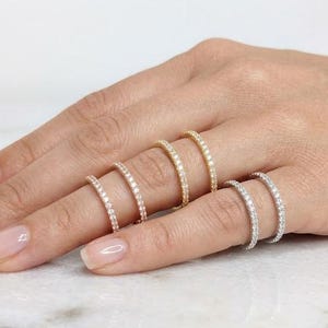 Puede incluir: Una mano muestra seis delicados anillos de eternidad en varios tonos metálicos: oro rosa, oro amarillo y plata. Cada anillo está adornado con pequeñas gemas brillantes, creando un efecto brillante. Los anillos se exhiben sobre una superficie clara.