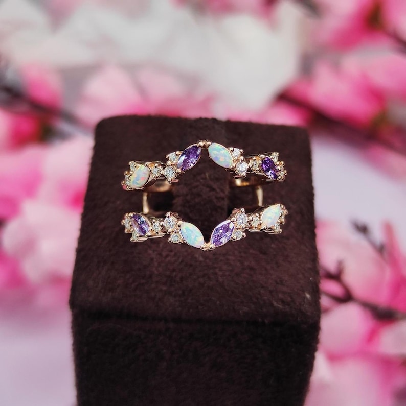 Op de afbeelding: Een goudkleurige ringversterker met een gebogen ontwerp. Het heeft afwisselende marquise-geslepen paarse en witte edelstenen, samen met kleine heldere accentstenen. De ring wordt weergegeven op een bruin oppervlak met een vage roze bloemenachtergrond.