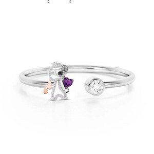 May include: A silver-toned open cuff bracelet featuring a stylized character holding a purple heart-shaped gem. The character has a silver body, black eyes, and a rose gold ear. A clear, round gem is set on the opposite end.